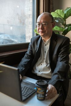 亚洲商人在一家舒适的咖啡馆里，对着窗户用笔记本电脑工作
