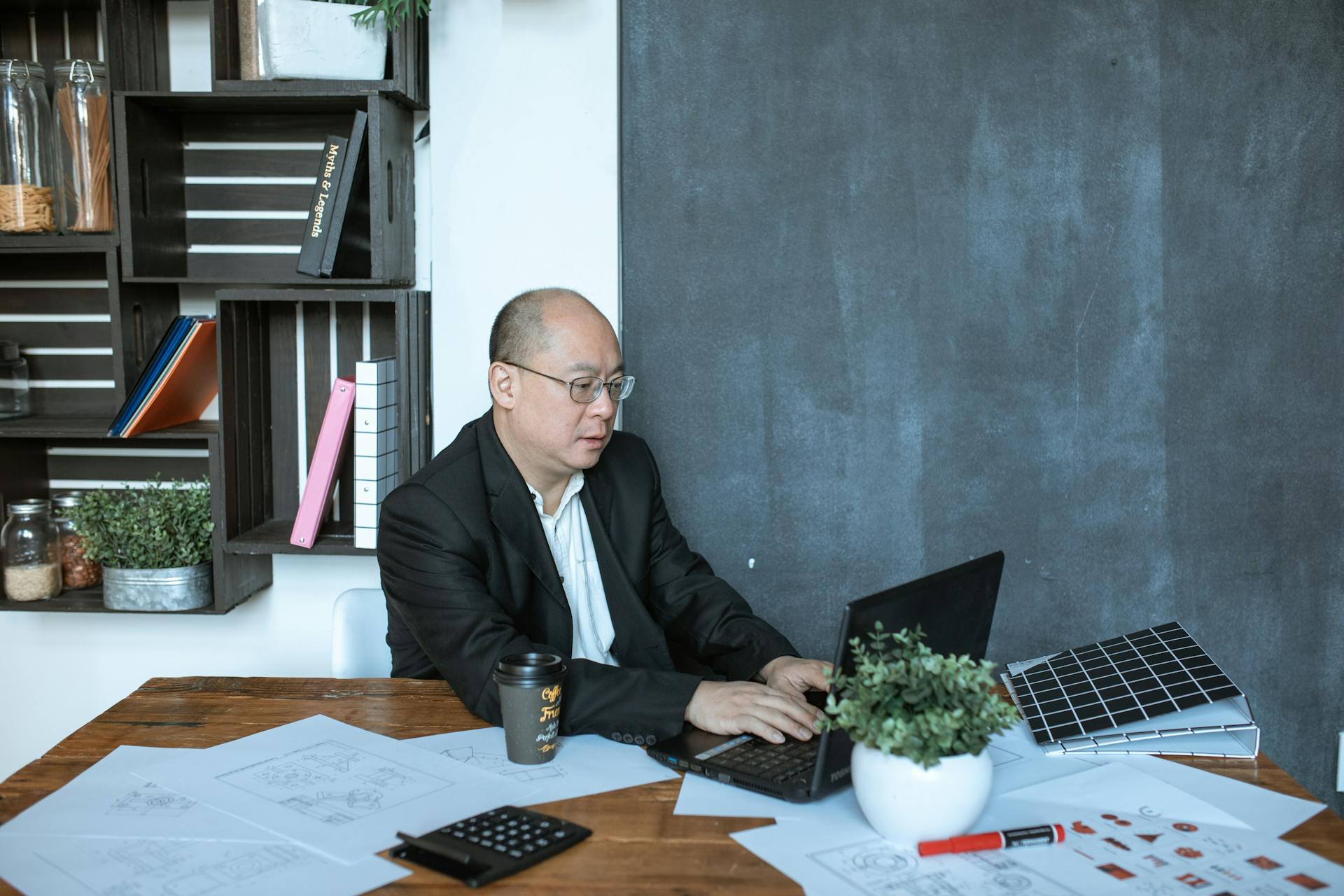 Asian businessman focused on laptop work in modern office with documents and coffee.