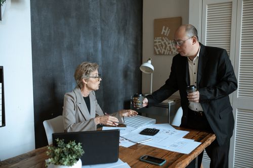 Free Two professionals in a modern office discussing work over coffee. Papers and laptop on desk. Stock Photo