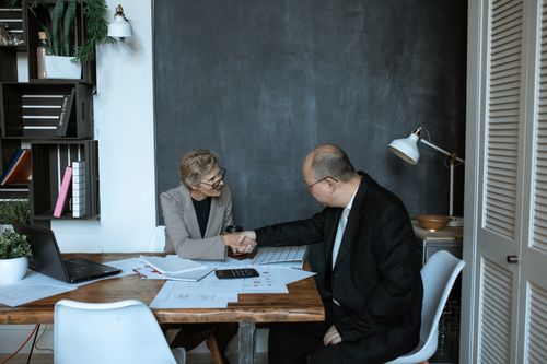 Free Two professionals engaged in a handshake across a desk in a modern office setting. Stock Photo