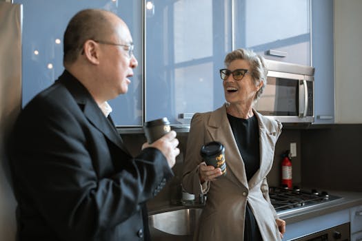 Two senior adults sharing a laugh over coffee in a modern office kitchen setting.