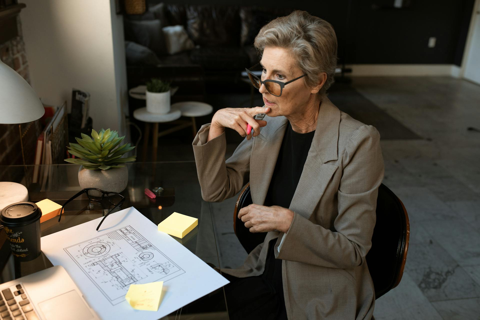 A senior architect in a gray blazer works thoughtfully at a home office with plans and a coffee.