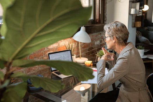 Senior woman in a modern home office focusing on work at a laptop next to a brick wall.