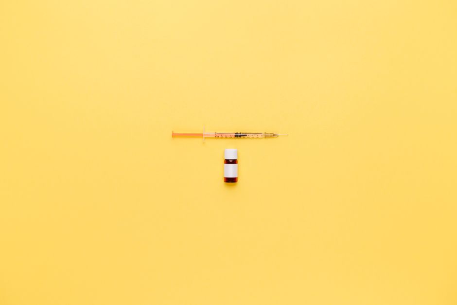 Minimalist image of a syringe and vaccine vial on a vibrant yellow background.