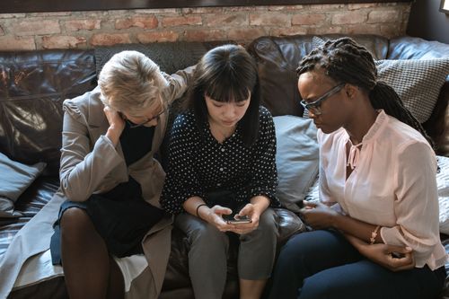 Free Three diverse women collaborating in a cozy office, fostering teamwork and creativity around a mobile device. Stock Photo
