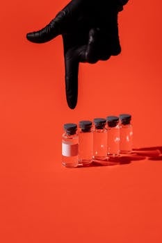 Black gloved hand pointing at a row of transparent vaccine vials on red background.