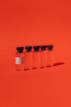 五个玻璃疫苗小瓶排列在明亮的红色表面上，投下强烈的阴影和高光