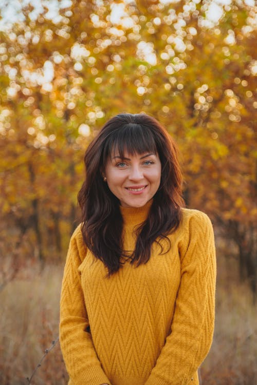 Cheerful dark haired female in sweater standing in autumn park and smiling while looking at camera