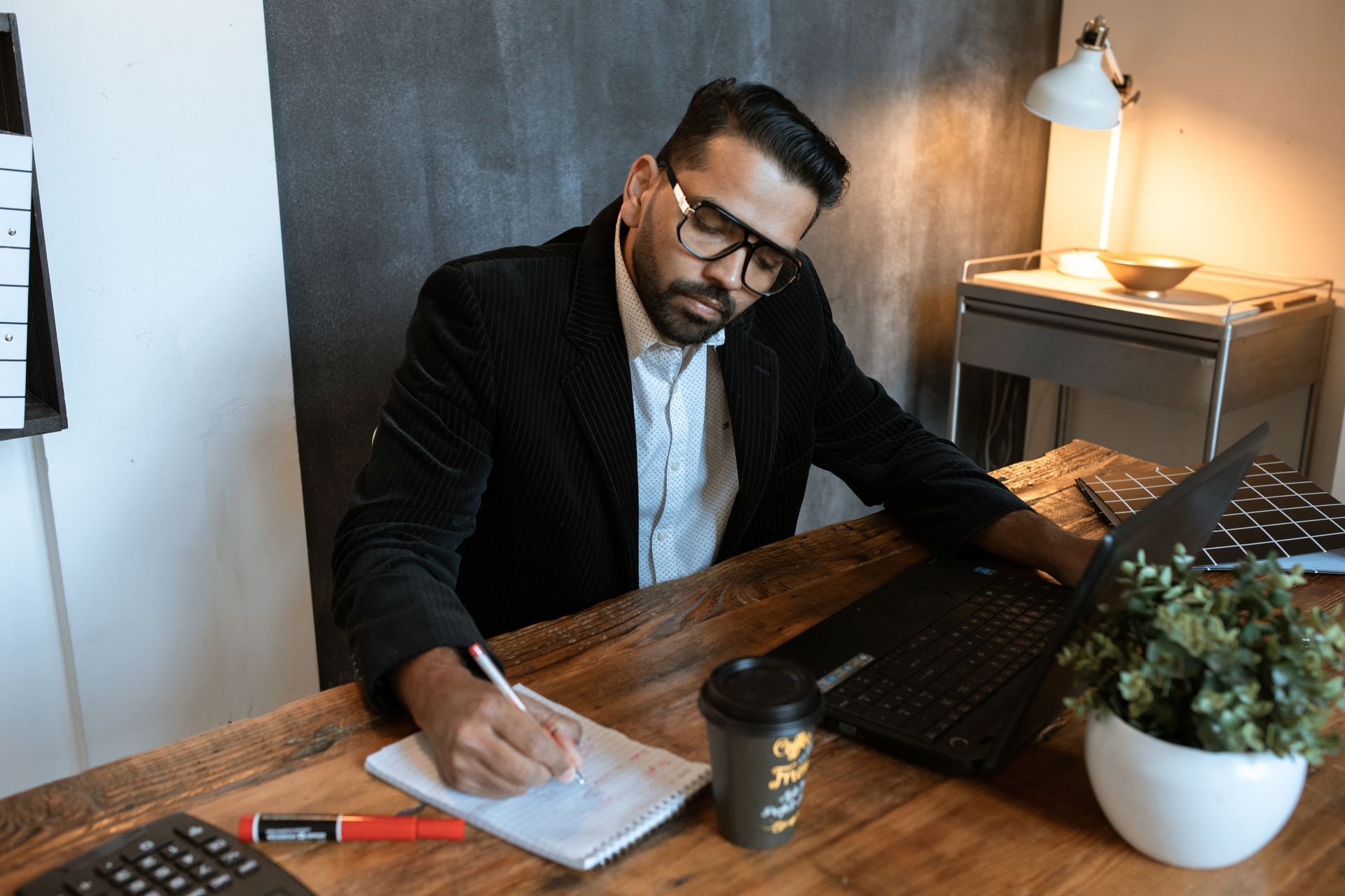A bearded man concentrating, writing notes, and using a laptop in a modern workspace.
