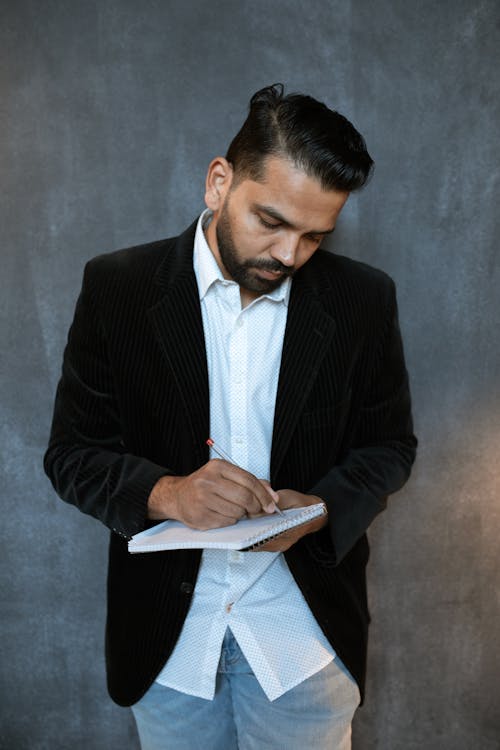 A Man in Black Suit Leaning on Gray Wall while Writing on a Notepad