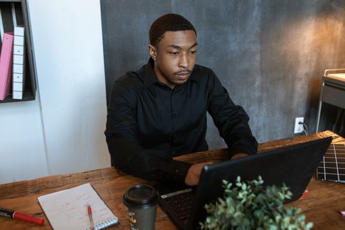 Free Man in office setting typing on laptop with coffee and notepad, showing concentration and productivity. Stock Photo