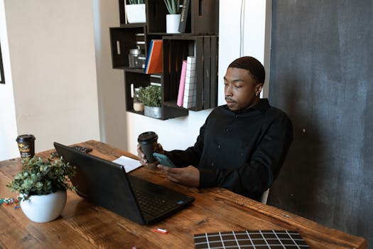 Black businessman working at a desk with laptop and phone in modern office setting.