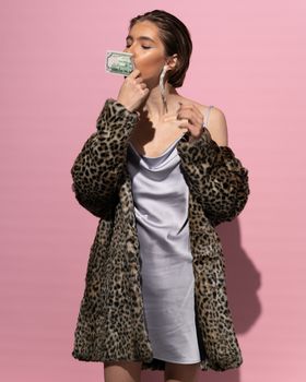 一位穿着皮草外套和丝绸连衣裙的时尚女性，手持现金，在粉色背景下散发着魅力