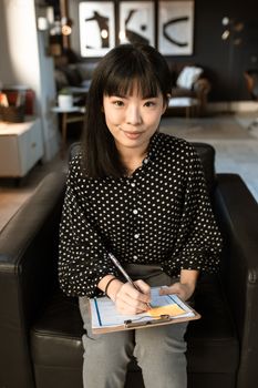 一位亚洲女性在舒适现代的客厅里，穿着波尔卡圆点上衣，正在剪贴板上写字
