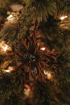 Closeup of wooden snowflake shaped decoration hanging on branch of lush Christmas tree with glowing garlands