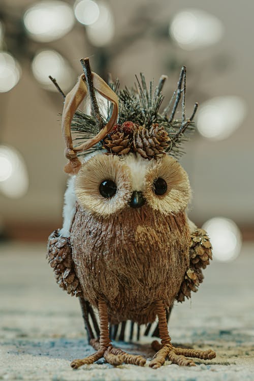 Statuette of little brown owl decorated with green twigs and pine cones placed on surface against blurred background with bokeh