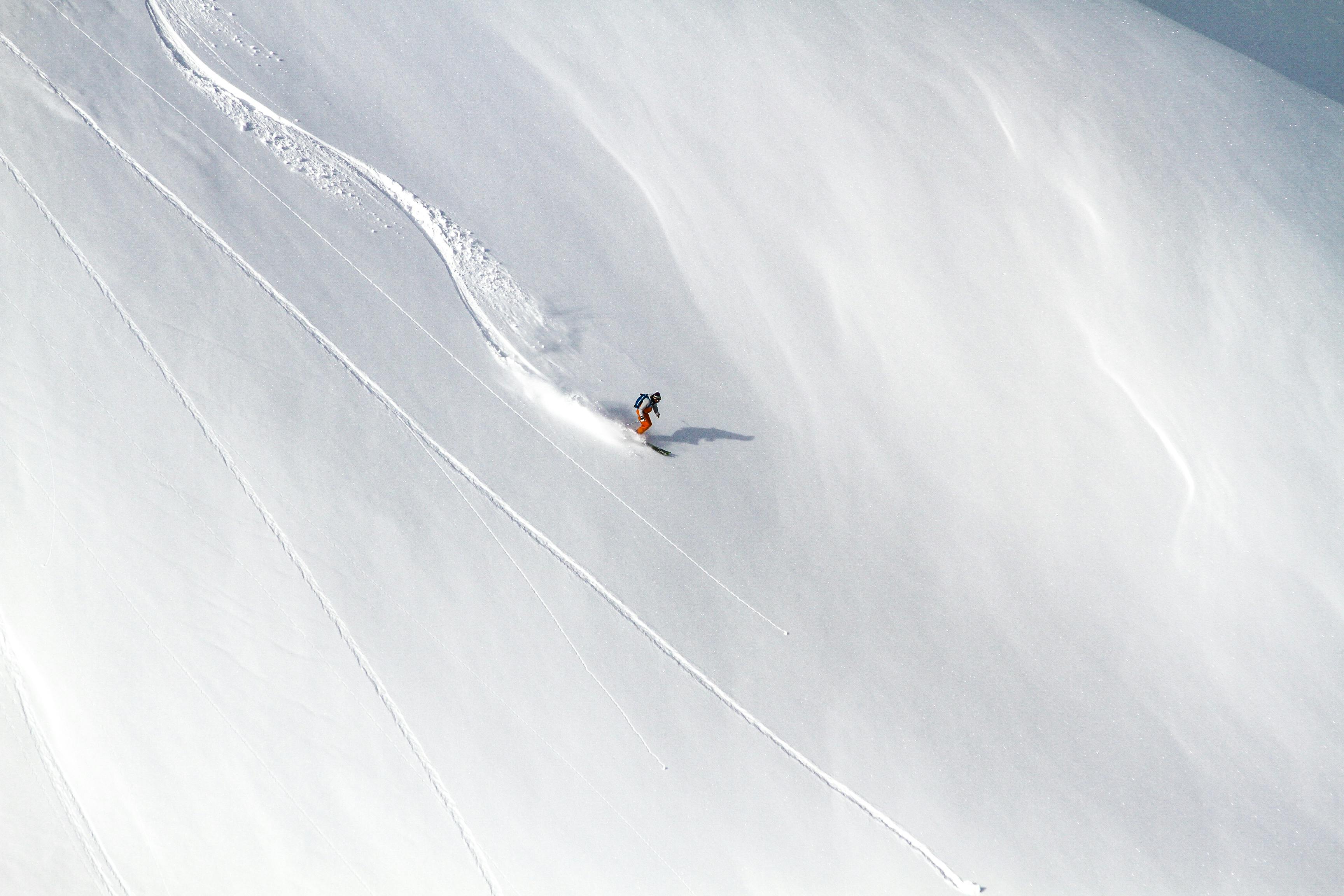 An aerial shot of a skier carving down a snow-covered mountain in winter, leaving a trail behind.