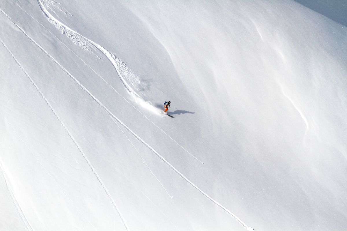 Aerial shot of a lone skier in an orange jacket carving through pristine white snow on a vast Swedish mountain, their track a single arc behind them