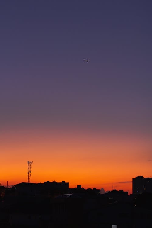 Colorful orange cloudless sky with crescent moon over dark residential buildings located on street in obscure city at sundown time