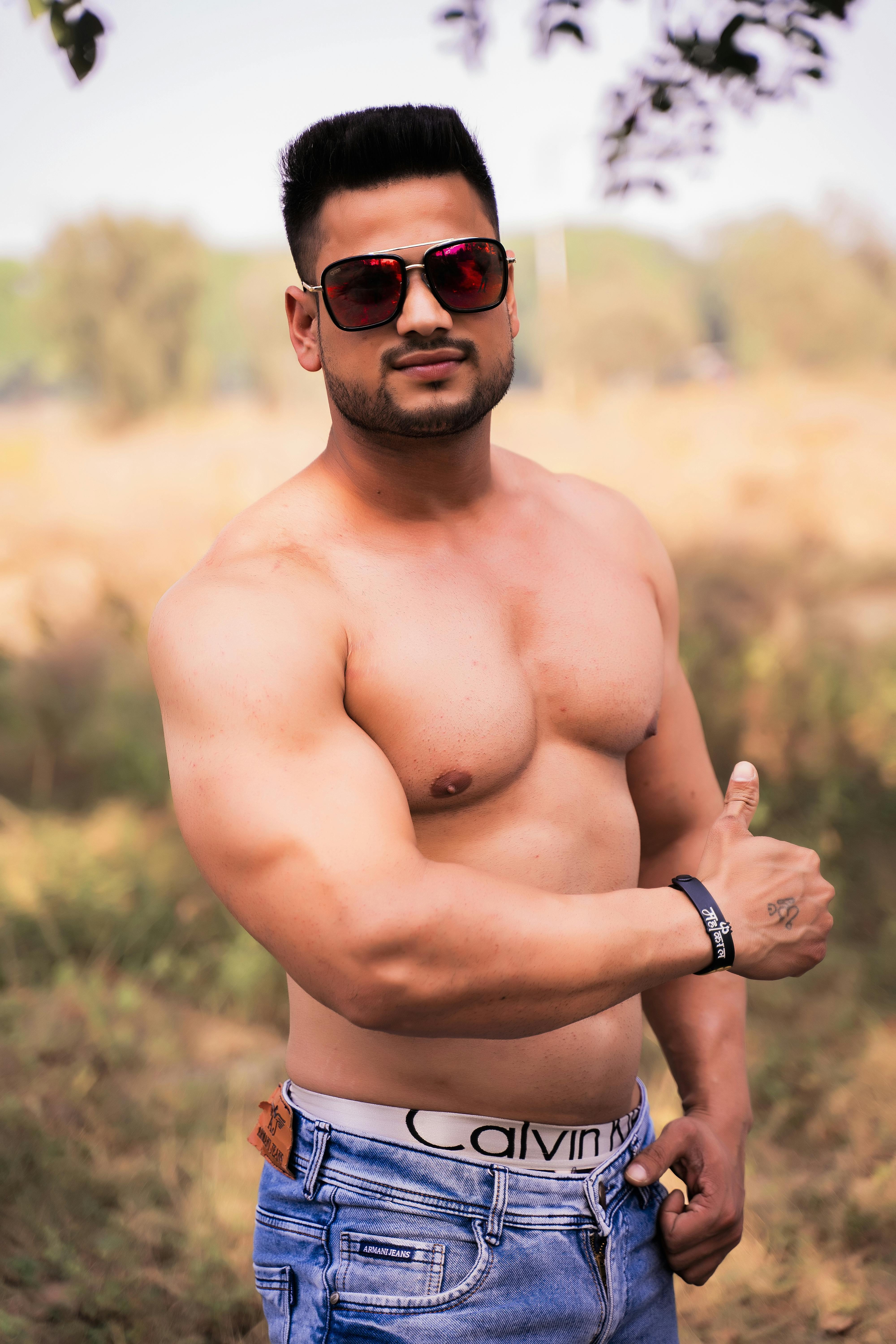 Portrait of a shirtless muscular man with sunglasses posing confidently outdoors during the day.
