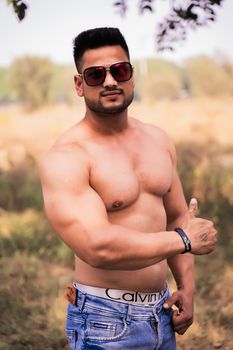 Portrait of a shirtless muscular man with sunglasses posing confidently outdoors during the day.