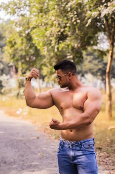 Shirtless muscular man flexing biceps in an outdoor nature setting, showing strength and fitness.