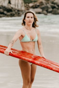 Young tattooed surfer in swimwear with surfboard looking at camera on ocean beach during summer trip