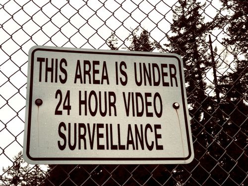 Free Sign on a chain-link fence stating 'This area is under 24 hour video surveillance.' Stock Photo