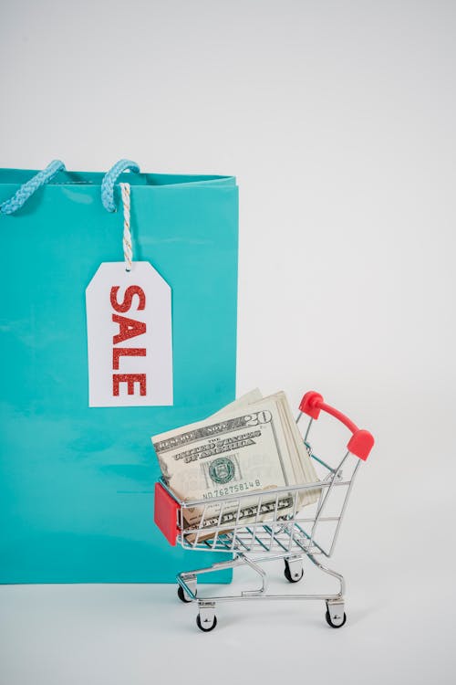 Shopping Bag With Sale Card Beside A Mini Push Cart With Dollar Bills