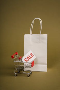 A miniature shopping cart with a red sale tag beside a white shopping bag on a neutral background, perfect for retail concepts.