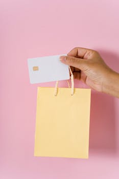 Close-up of a hand holding a credit card and yellow shopping bag on a pink backdrop, symbolizing online shopping.