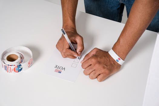 A person wearing a wristband fills out a voting ballot with a pen. Voting stickers visible.