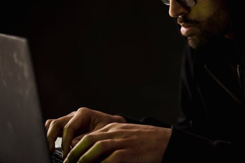 Free Crop concentrated bearded male in eyeglasses and black clothes browsing modern netbook in dark room Stock Photo