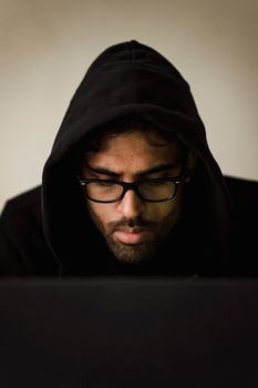 一名戴着眼镜和兜帽的男子，在昏暗的光线下专注地操作着笔记本电脑