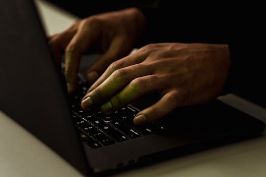 A close-up view of hands typing on a laptop keyboard in a dimly lit environment.