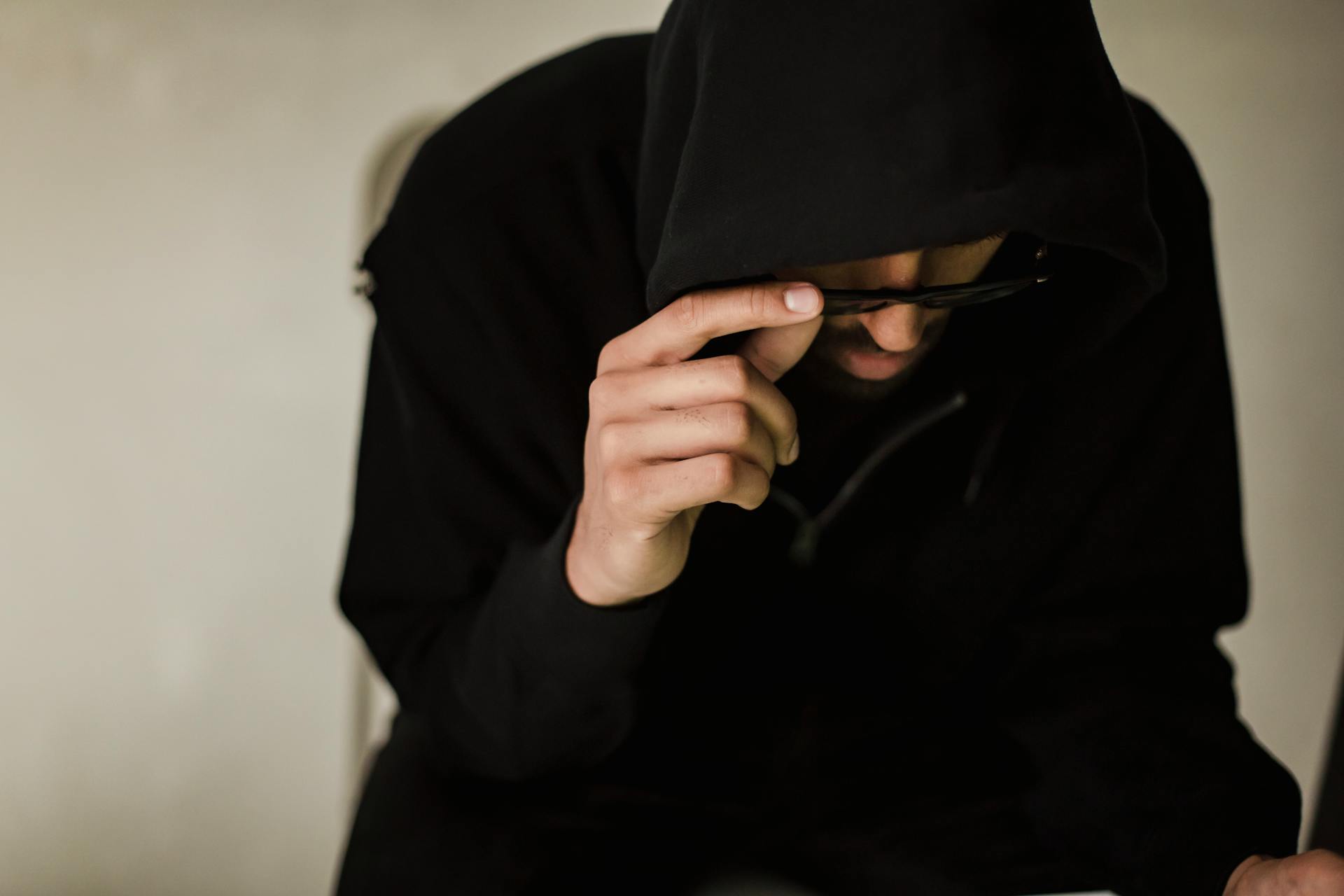 A man in a dark hoodie looks pensive while using a device indoors, symbolizing cyber themes.