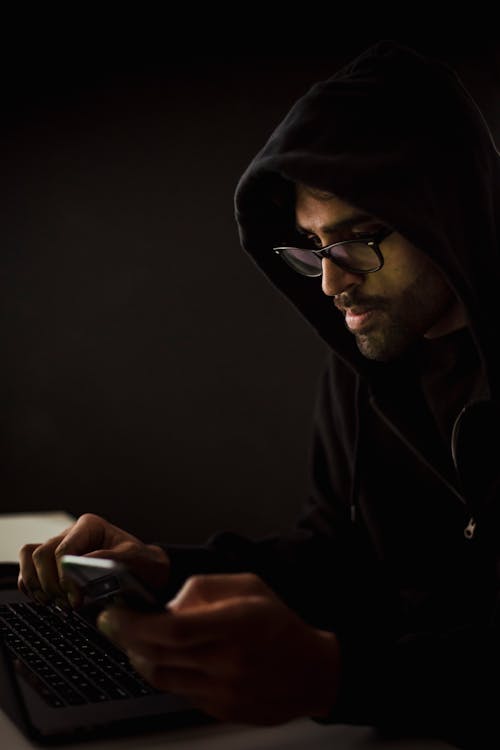 Free Side view concentrated male hacker in black hood browsing netbook and mobile phone while surfing dark net in obscurity Stock Photo