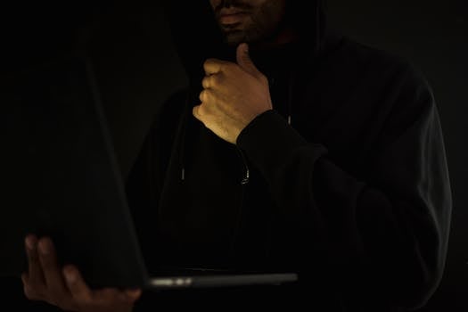A man in a hoodie uses a laptop in darkness, symbolizing cybercrime and anonymity.