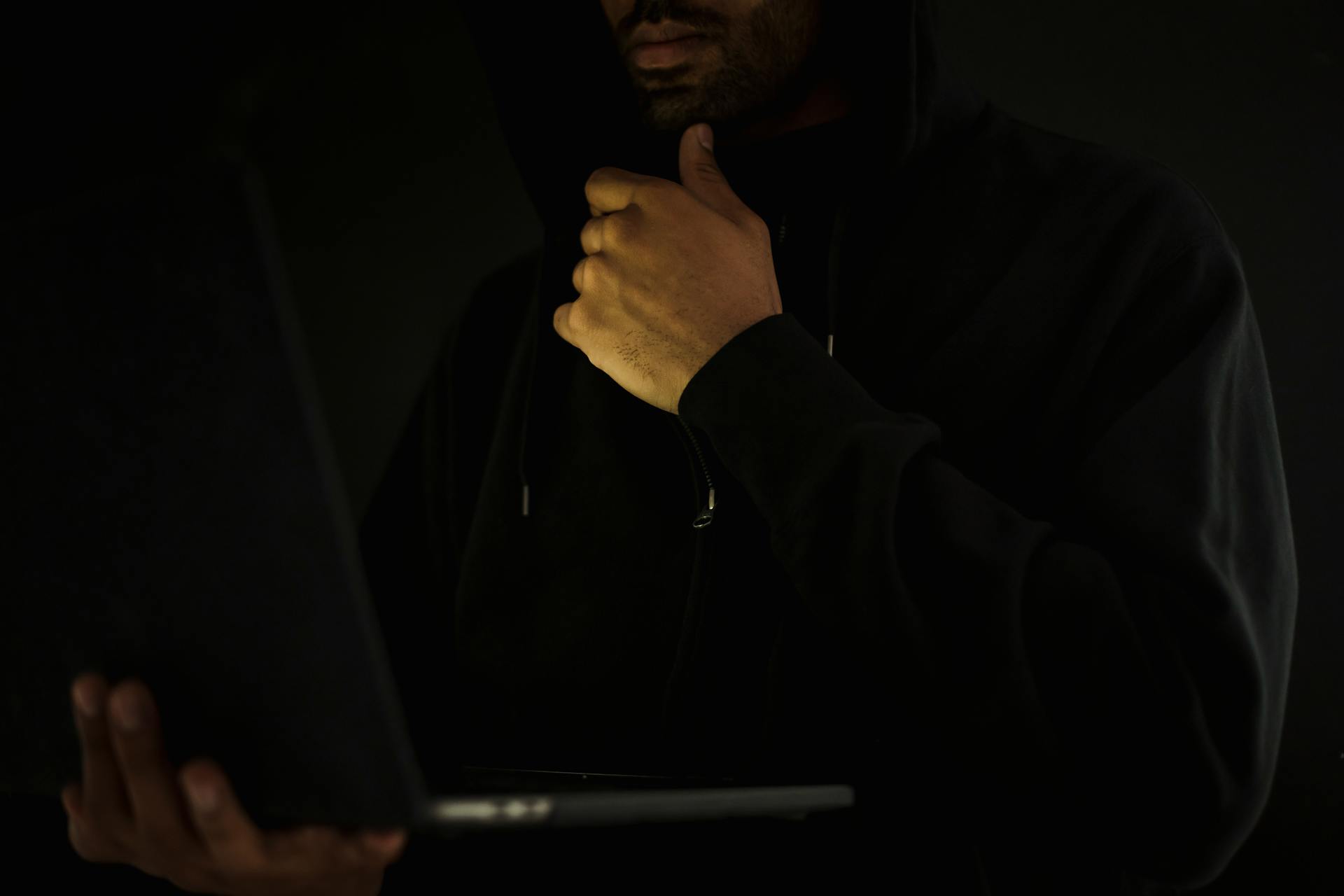 A man in a hoodie uses a laptop in darkness, symbolizing cybercrime and anonymity.