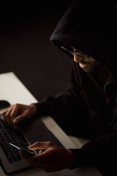 From above crop focused male hacker in black hood working on netbook and mobile phone while surfing cyberspace in dark room