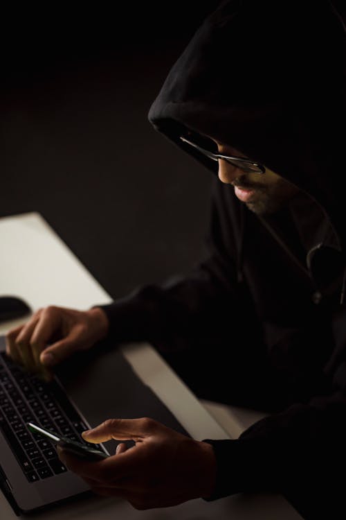 Free From above crop focused male hacker in black hood working on netbook and mobile phone while surfing cyberspace in dark room Stock Photo