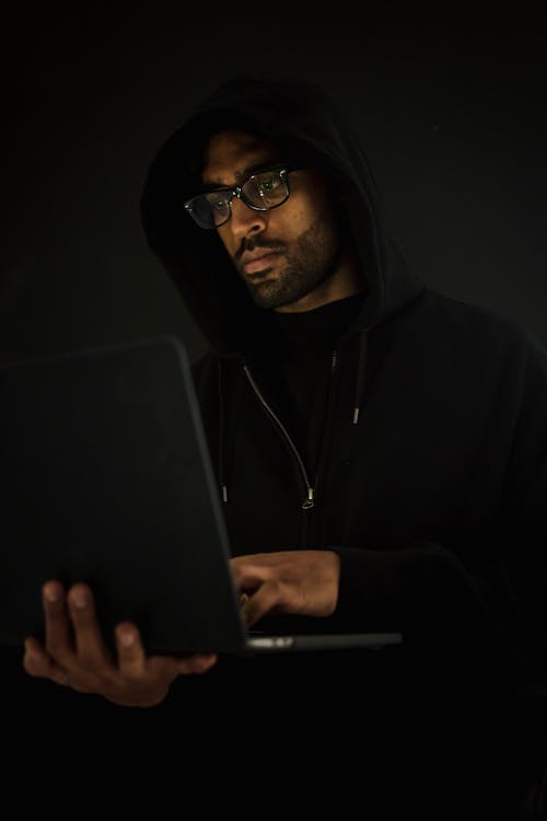 Concentrated bearded male hacker in black hoodie and eyeglasses holding netbook on hand and surfing dark net in obscurity
