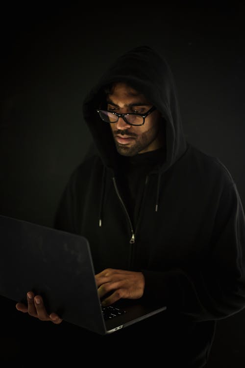 Free Serious young male in black hood and eyeglasses standing with netbook in hand and hacking database in dark room Stock Photo