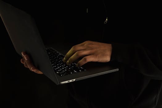Silhouette of a person using a laptop in a dark environment, emphasizing online work or security.
