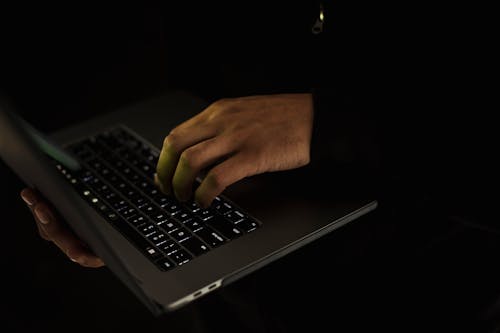 Free Crop unrecognizable male in black sweater holding netbook on hand and typing on keypad in dark room Stock Photo