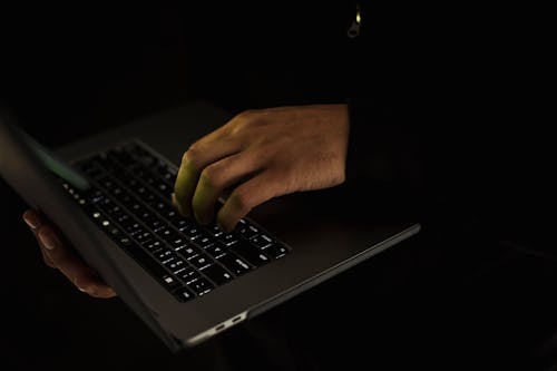 Crop unrecognizable male in black sweater holding netbook on hand and typing on keypad in dark room