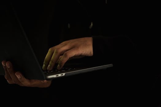 A close-up of a person's hands typing on a laptop in a dark setting, suggesting secrecy or privacy.