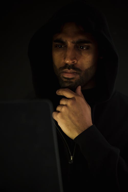 Free Concentrated bearded male hacker wearing black hood looking at netbook screen and touching chin in contemplation while hacking software in darkness Stock Photo