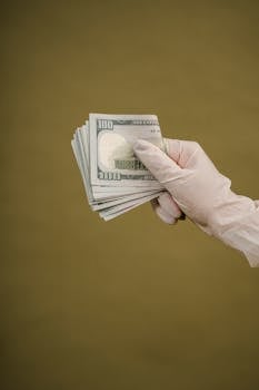 A gloved hand holding a bundle of dollar bills against a neutral background.