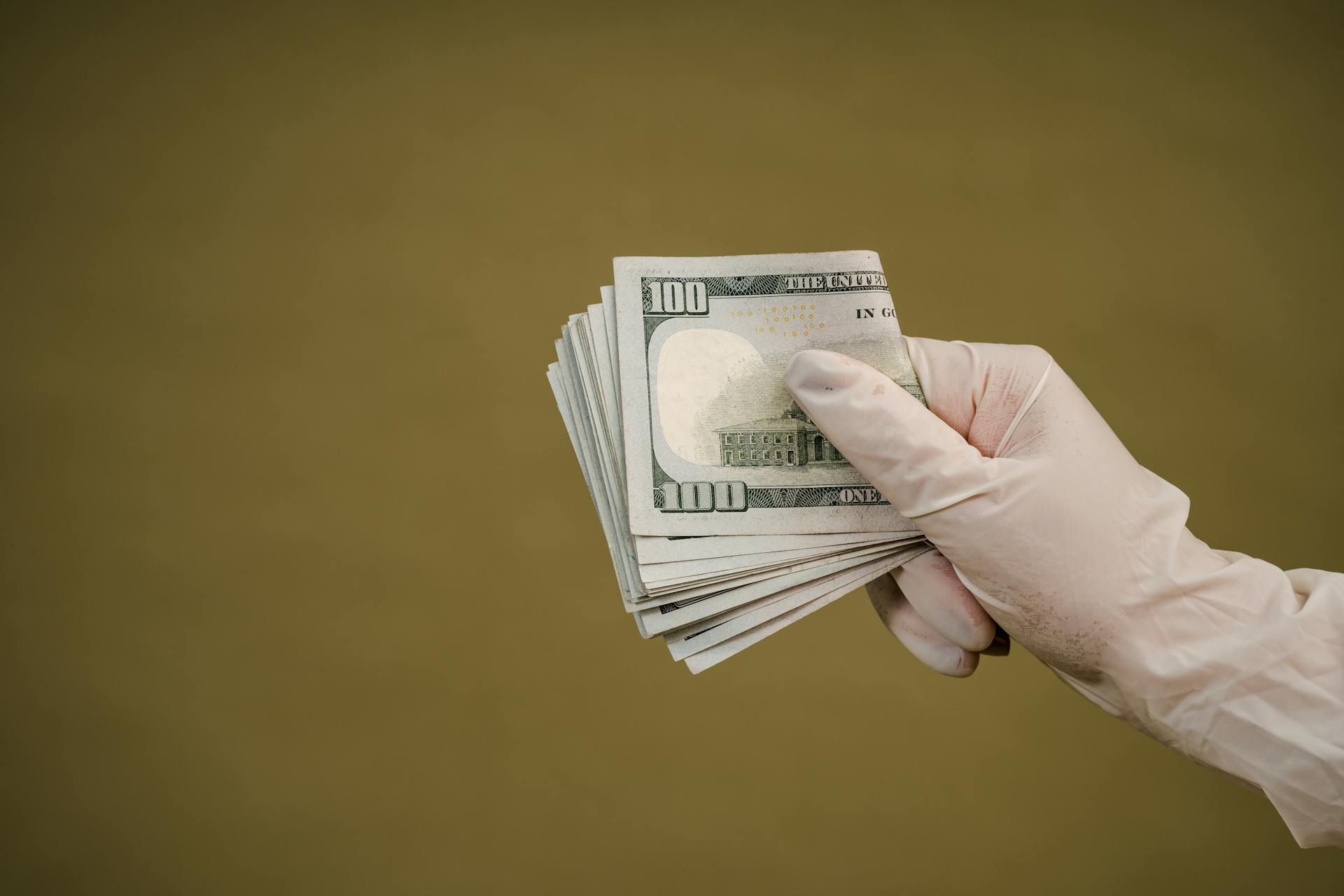 A gloved hand holds a stack of hundred-dollar bills against a plain background.